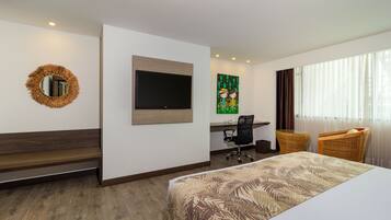 A hotel room with a bed, a desk with a chair, a television, a wicker chair, and a decorative mirror.