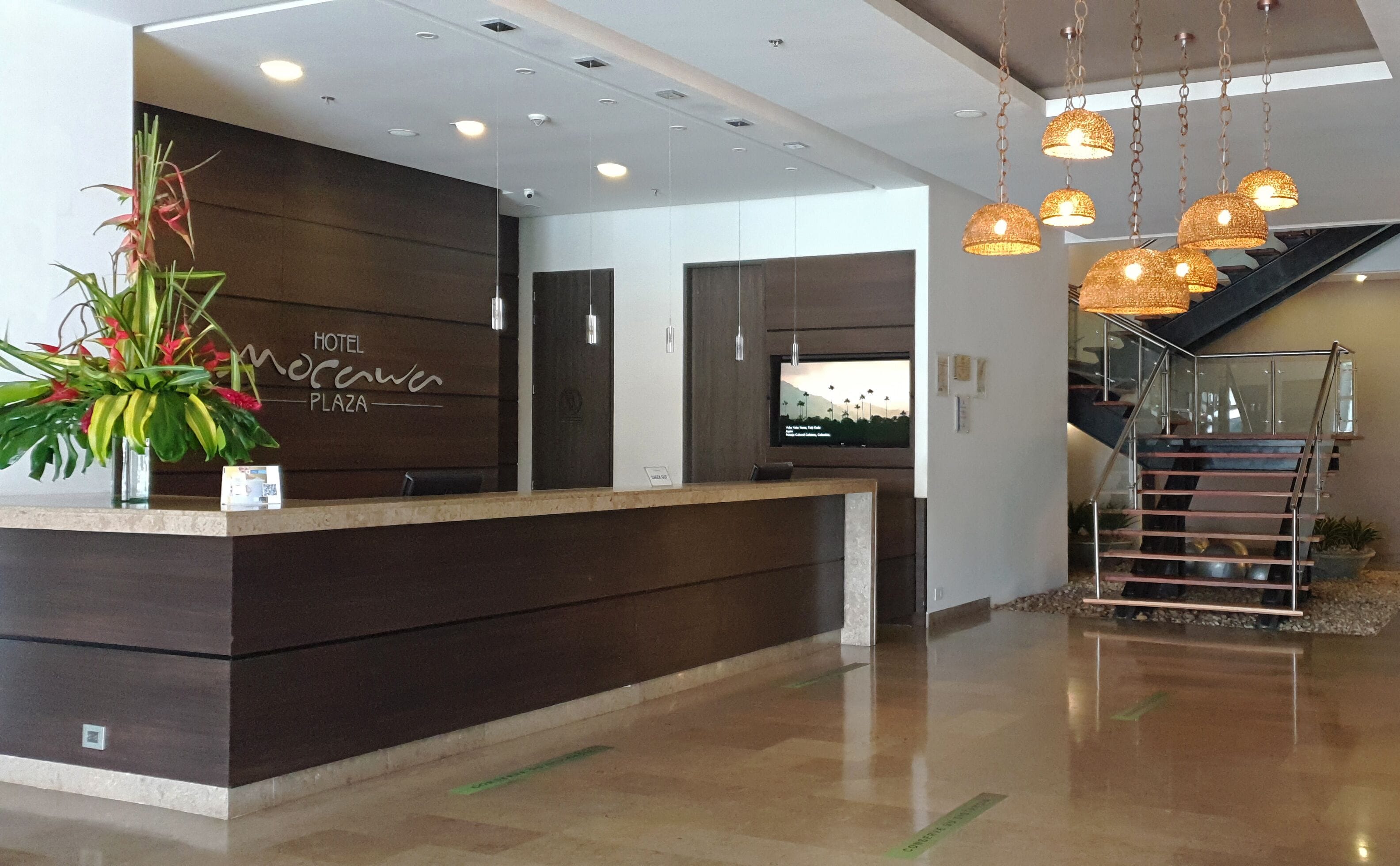 A hotel reception desk with a modern design, a large screen displaying a scenic view, and a staircase leading to upper floors.