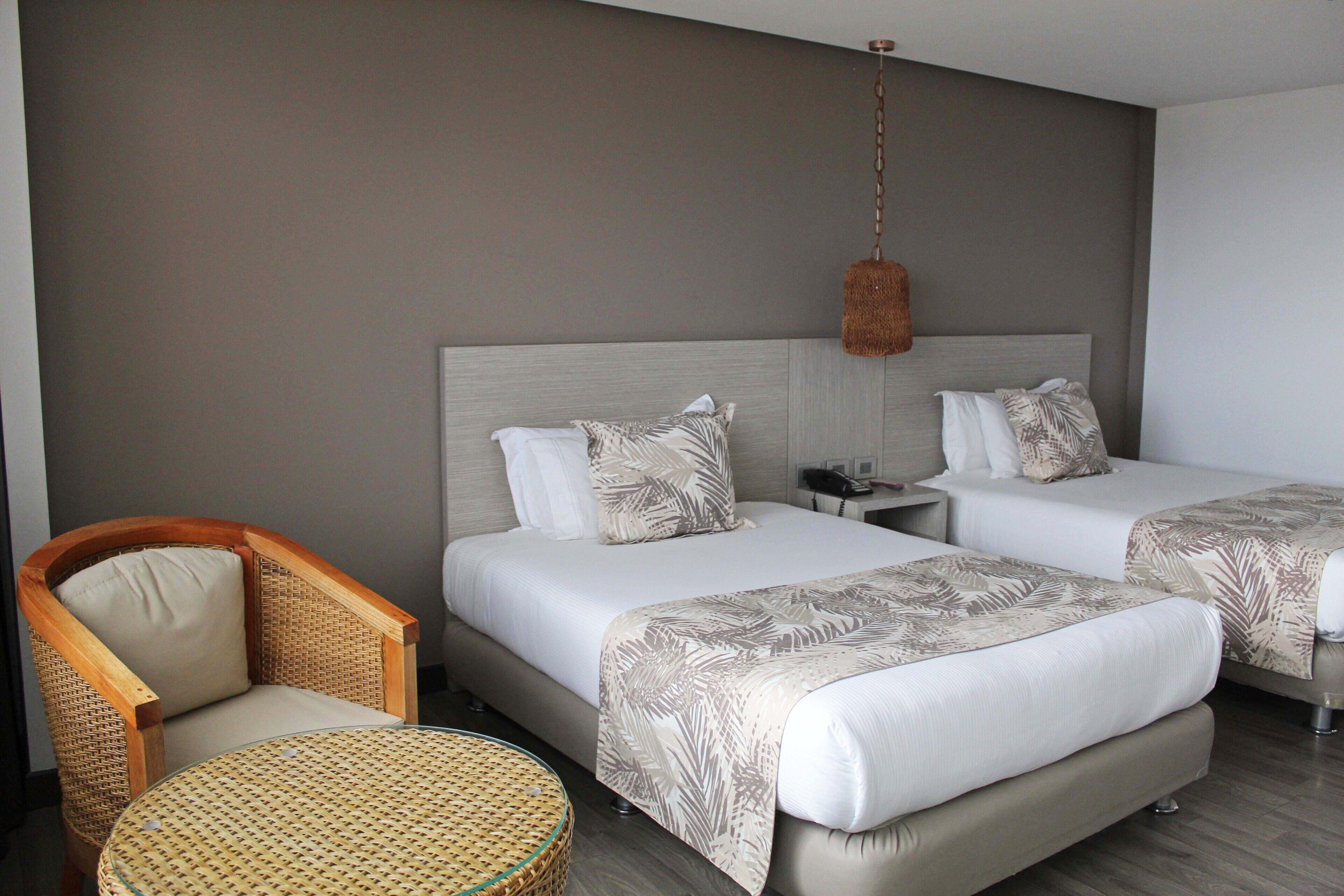 Habitación de hotel con dos camas, una silla de mimbre, una mesita redonda y una lámpara colgante.
