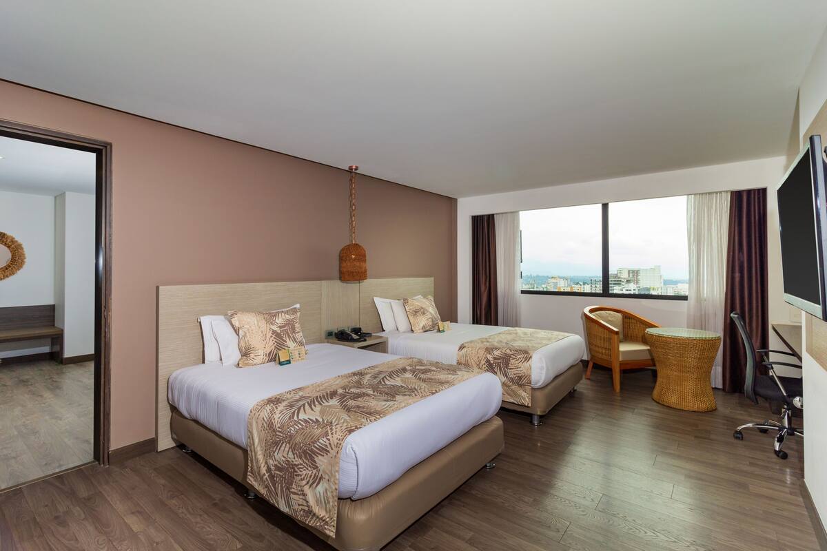 Habitación de hotel con dos camas, una silla, una mesita, televisor y vista a la ciudad.