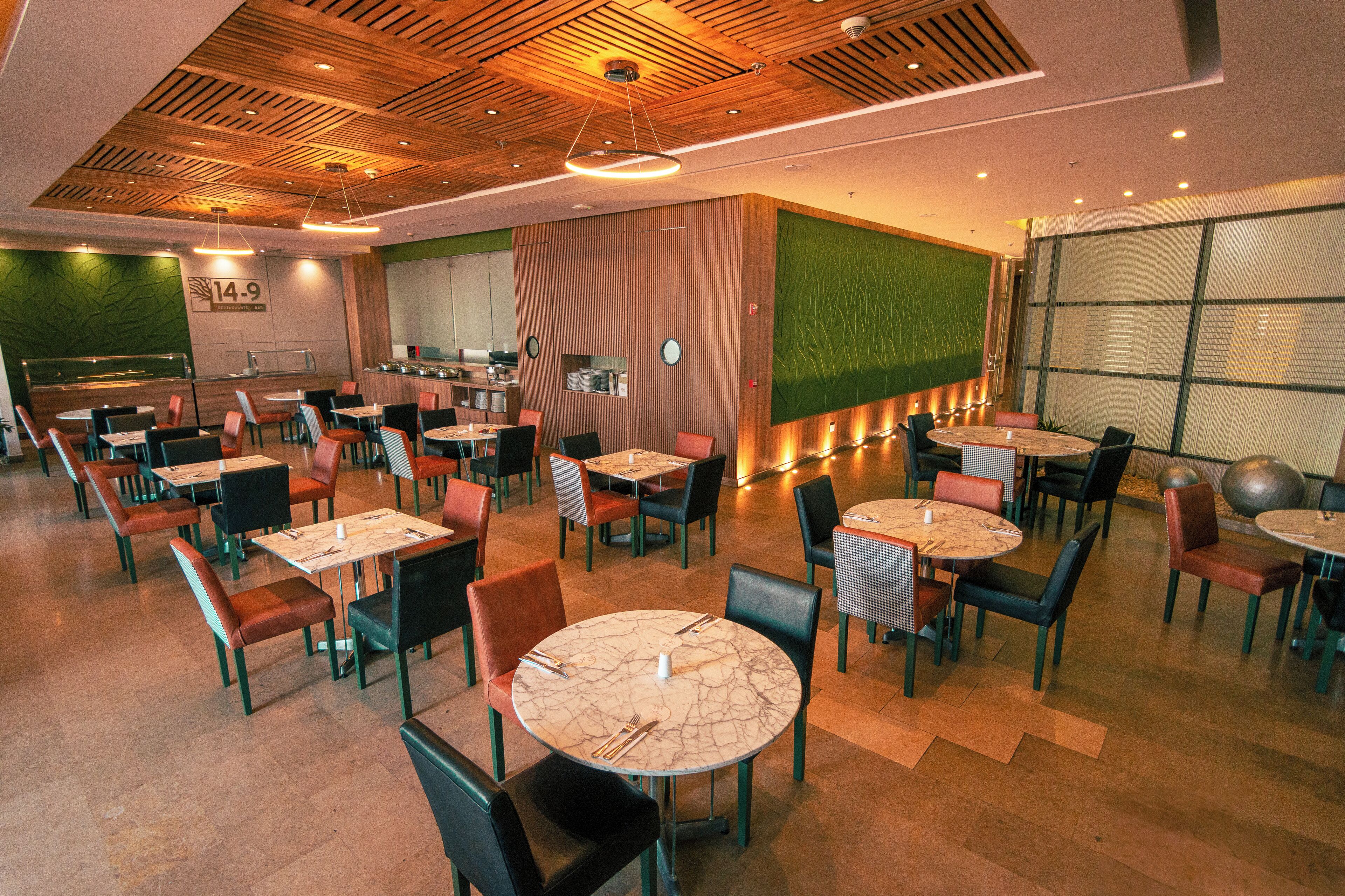 A modern restaurant with round tables and chairs, a wooden ceiling, and a green accent wall.