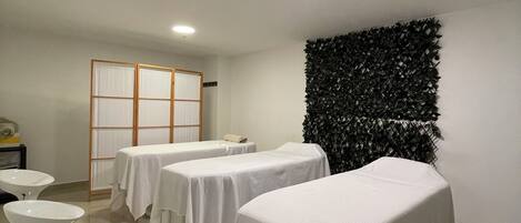 Una stanza con quattro lettini da massaggio ricoperti di lenzuola bianche, un pannello decorativo nero a parete e una porta scorrevole in legno.
