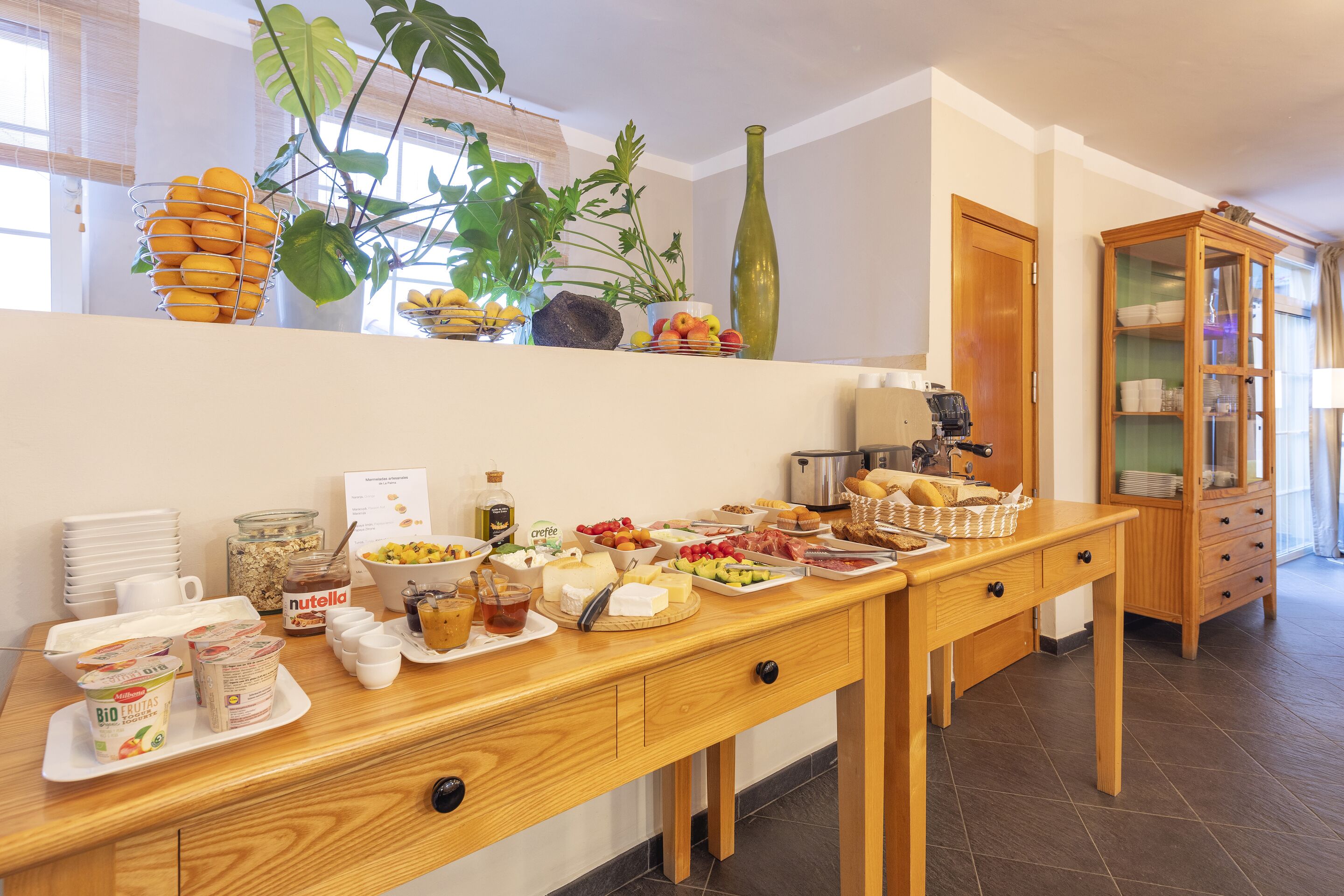A breakfast buffet with various food items and beverages, a wooden cabinet, and a plant.