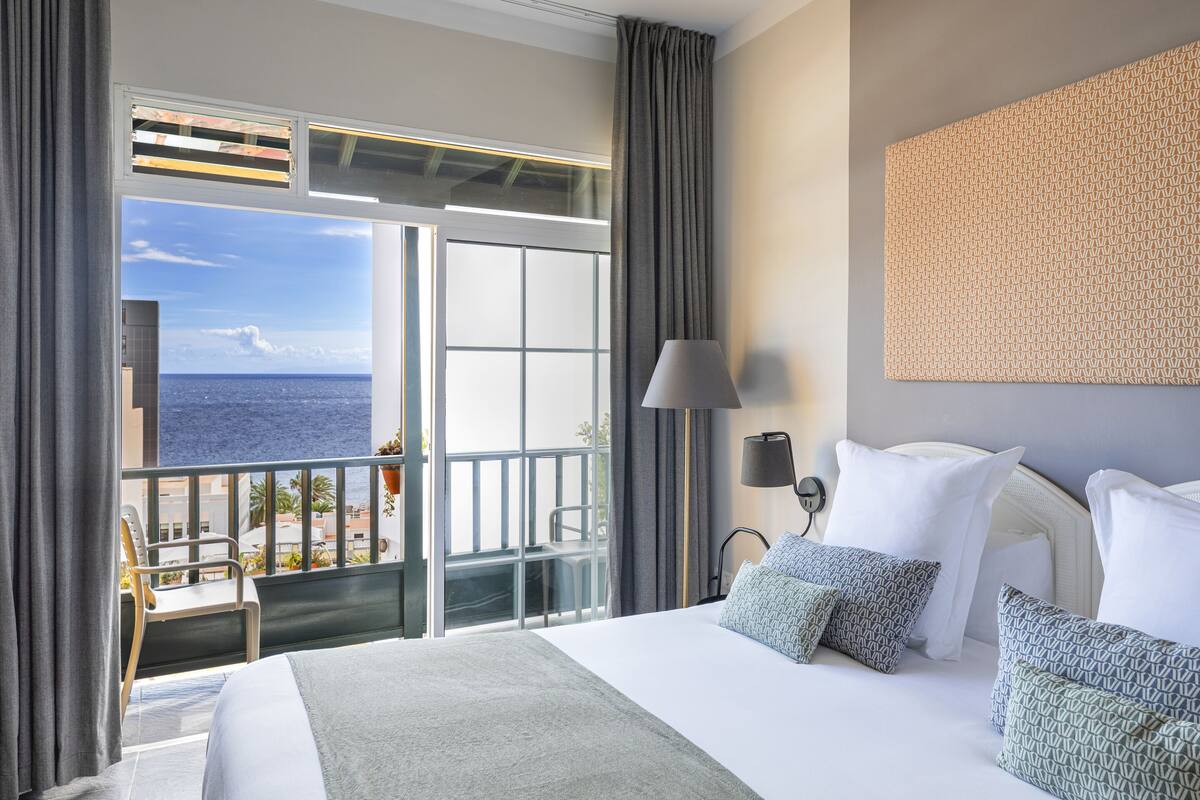 Hotelkamer met een groot bed, uitzicht op zee, een balkon met stoel en een behang met patroon.