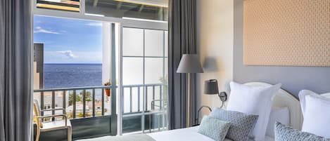 A hotel room with a large bed, a view of the sea, a balcony with a chair, and a patterned wall.