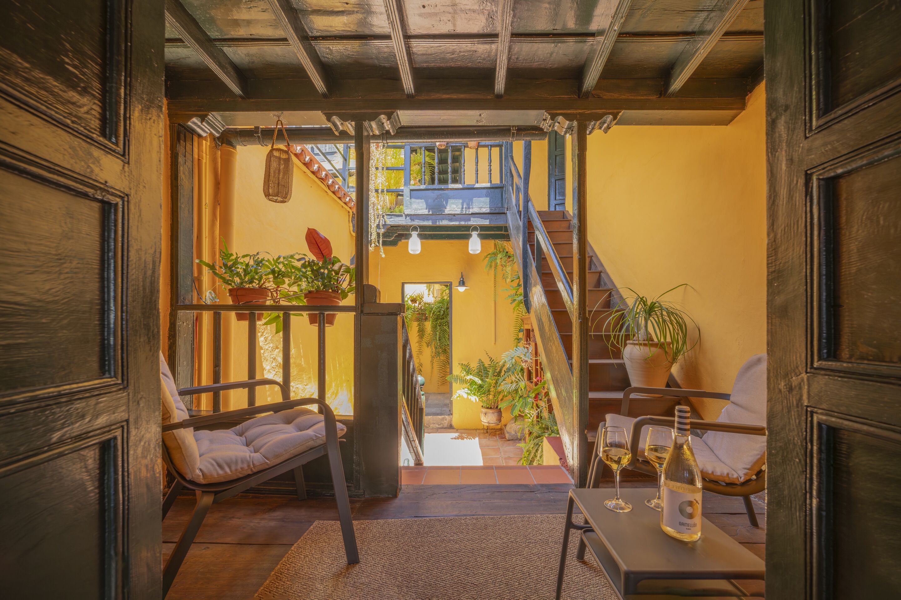 Un interior acogedor con una escalera que lleva a un balcón, plantas en macetas y una mesa con una botella y vasos.