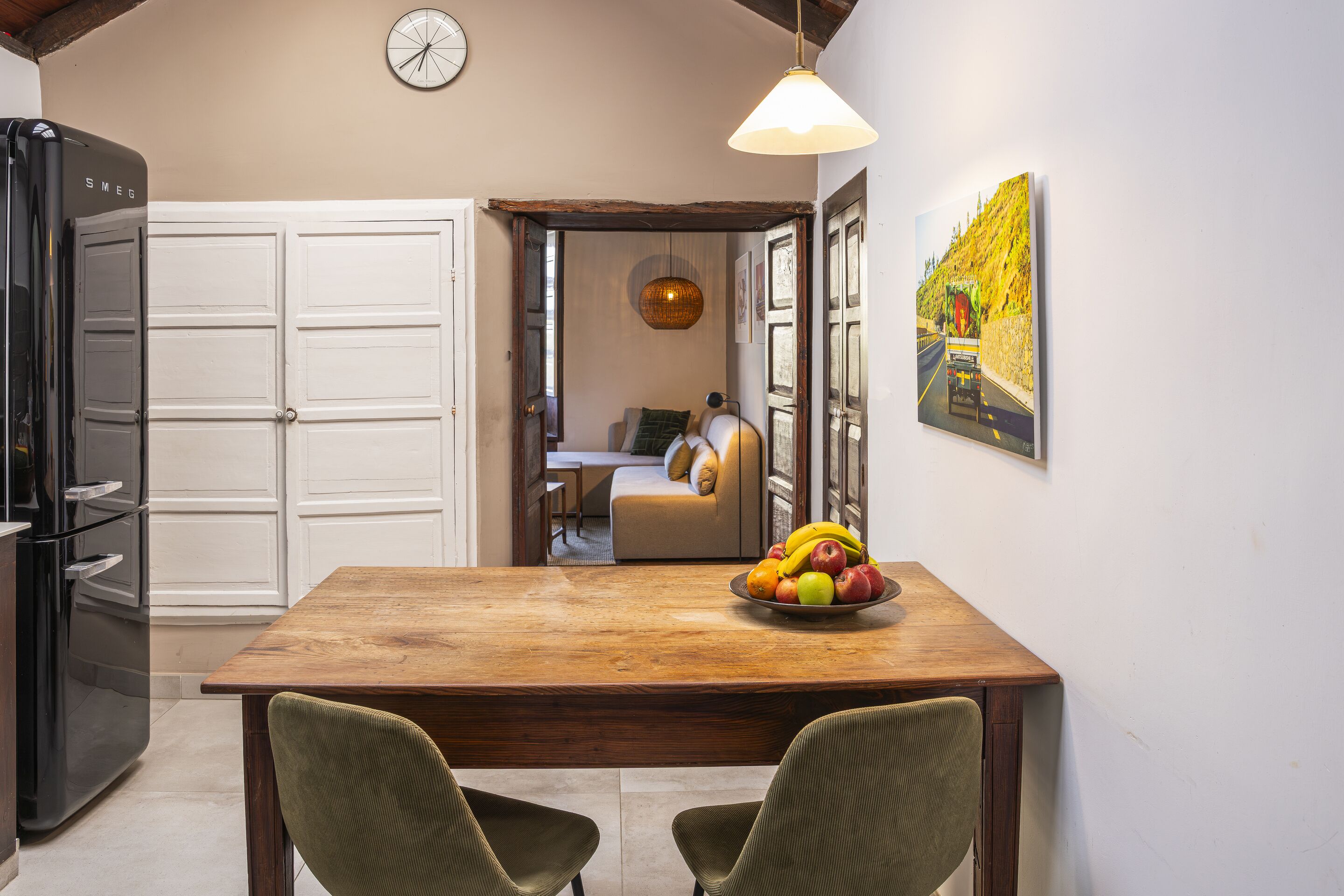 A dining area with a wooden table, two chairs, a fruit bowl, a clock, and a painting on the wall.