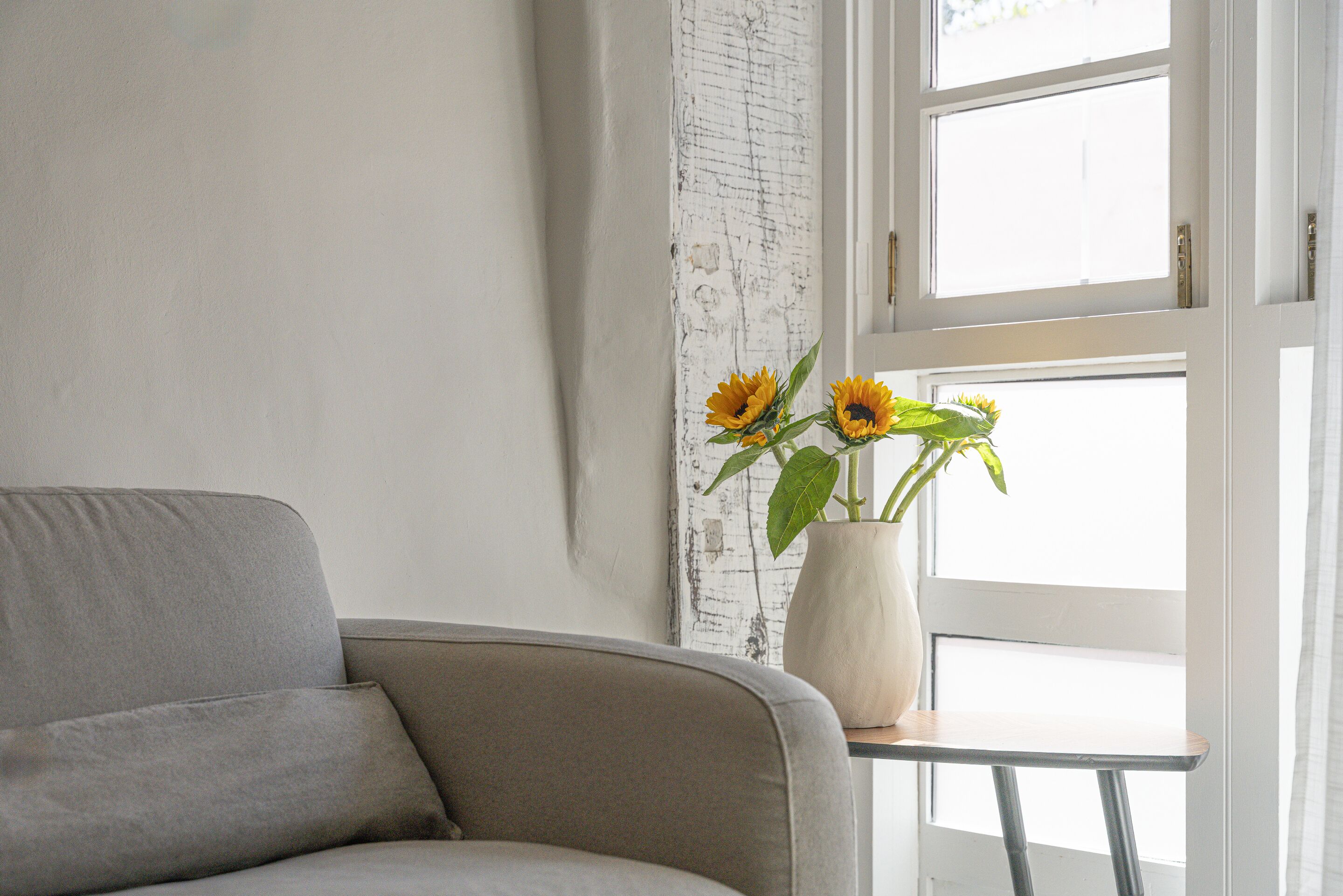 A room with a grey sofa, a small table with a vase of sunflowers, and a white wall with a textured artwork.