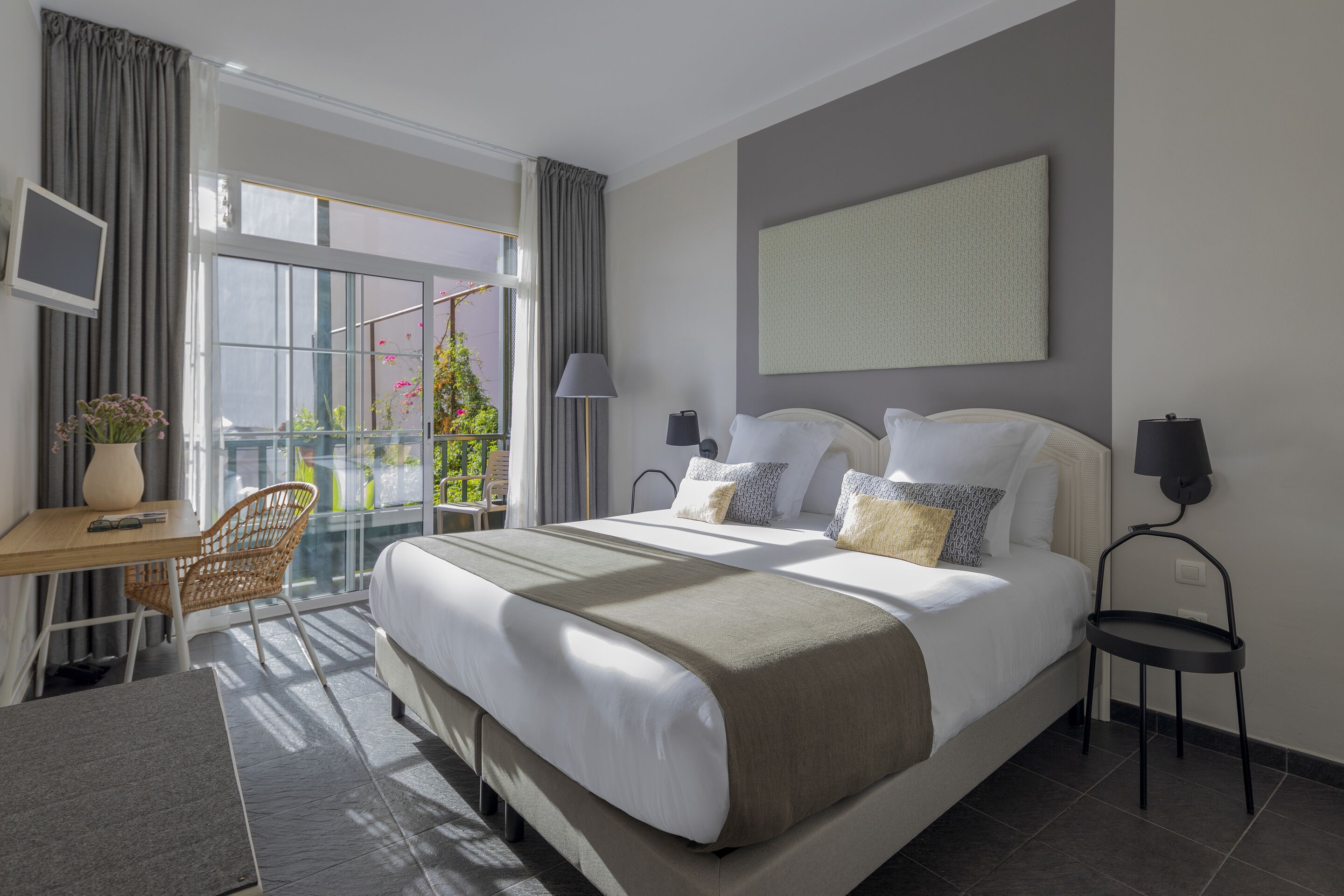 Une chambre d’hôtel moderne avec un grand lit, un bureau et une chaise. Elle dispose d’un balcon avec vue sur la verdure et d’une fenêtre avec des rideaux.