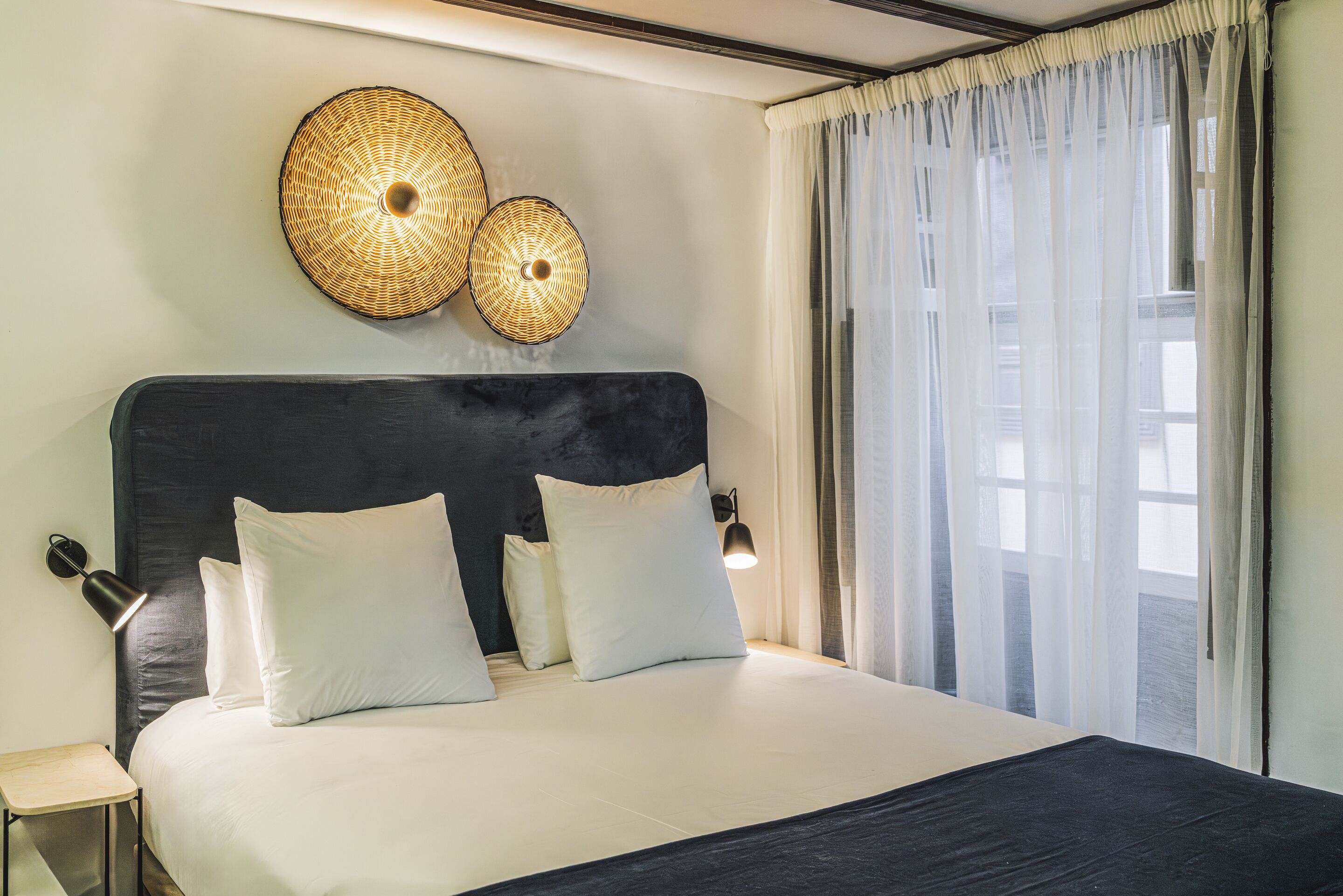 Una cama bien hecha con ropa de cama blanca y negra, un cabecero oscuro y dos lámparas fijadas a la pared.