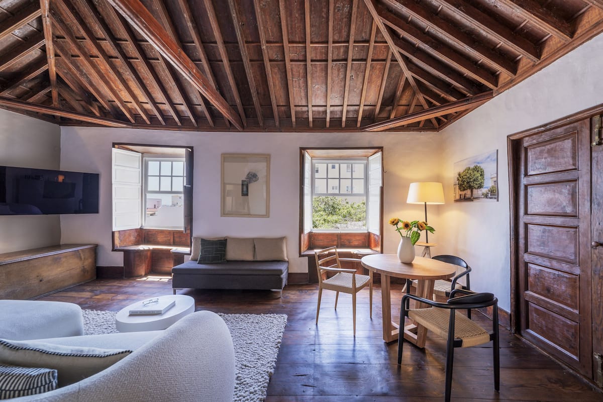 Een gezellige woonkamer met een houten plafond, een bank, een salontafel en een eethoek met stoelen en een lamp.