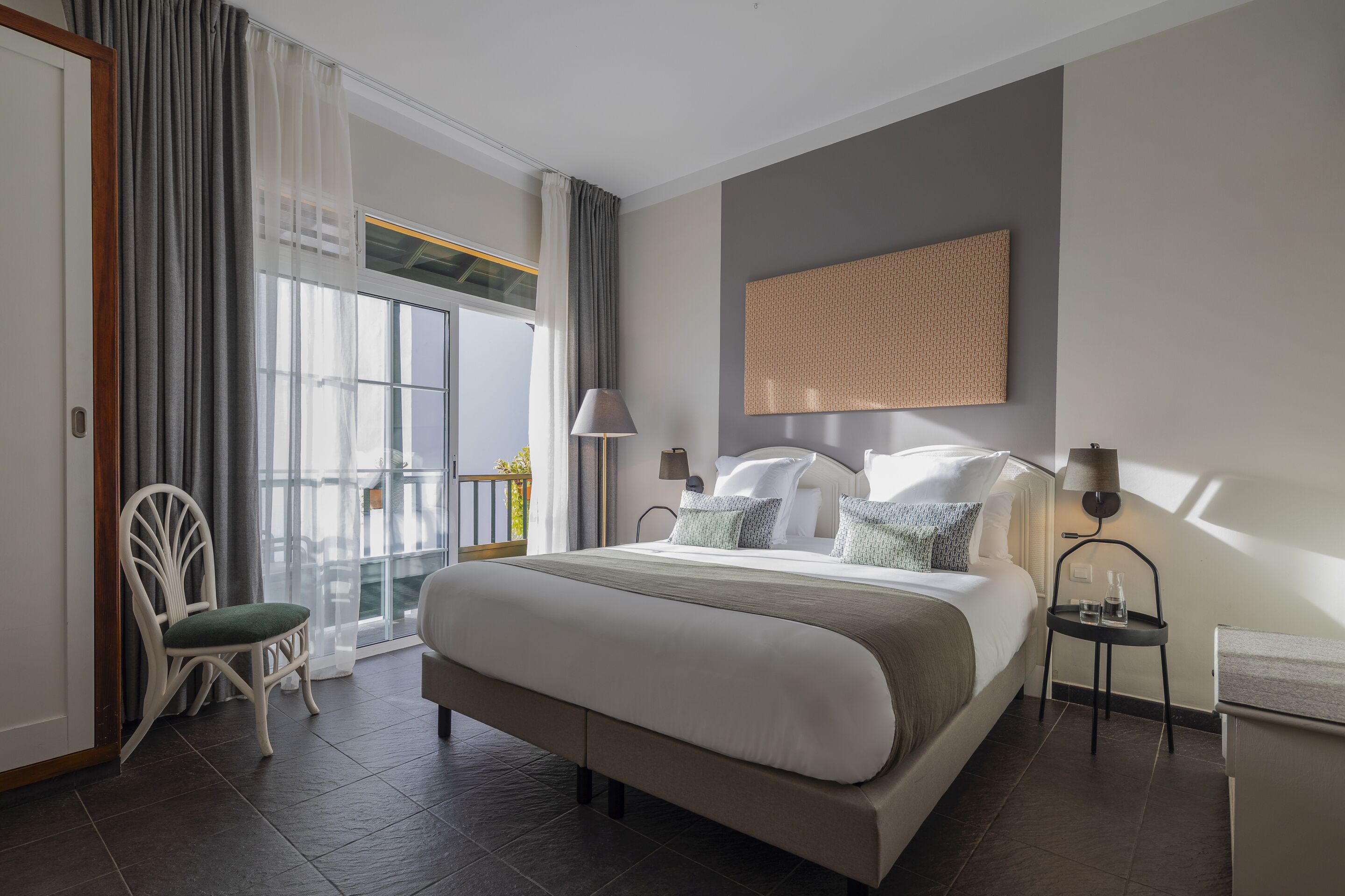 A modern hotel room with a large bed, a chair, a small table, and a view of the outdoors.