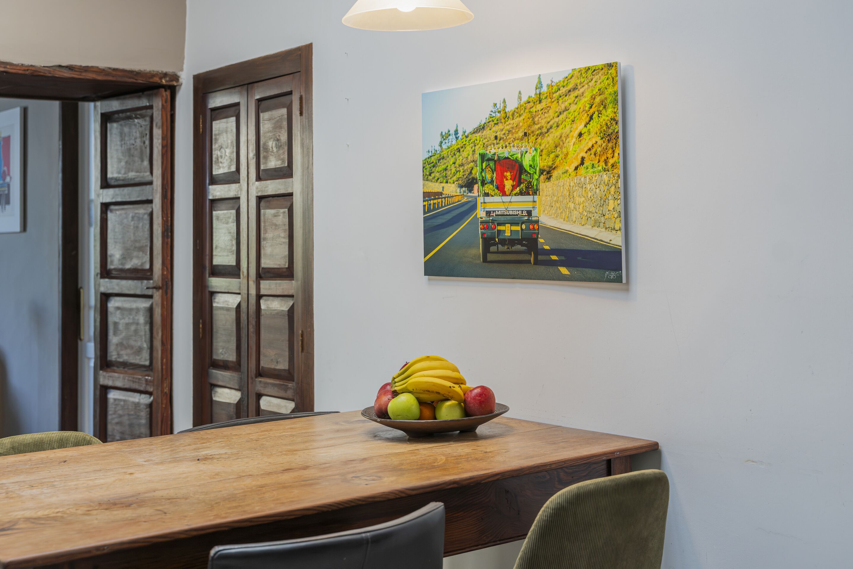 Un coin repas avec une table en bois, un saladier rempli de fruits et un tableau accroché au mur représentant un véhicule sur une route.