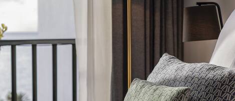A neatly made bed with patterned pillows, a pair of glasses, and a floor lamp in a hotel room with a view of the sea.