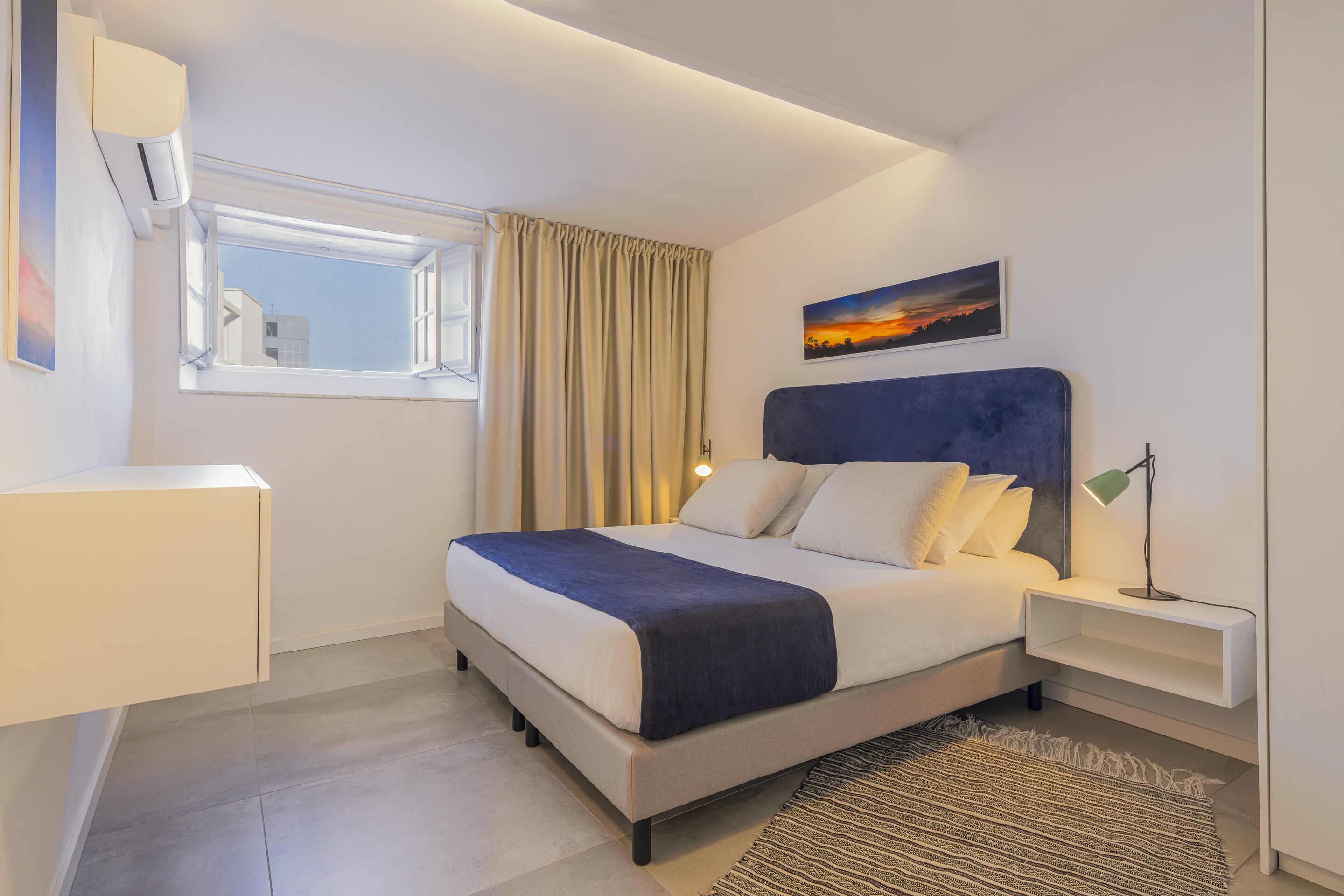A modern bedroom with a bed, bedside tables, a nightstand, a window with curtains, a painting on the wall, and a ceiling light.