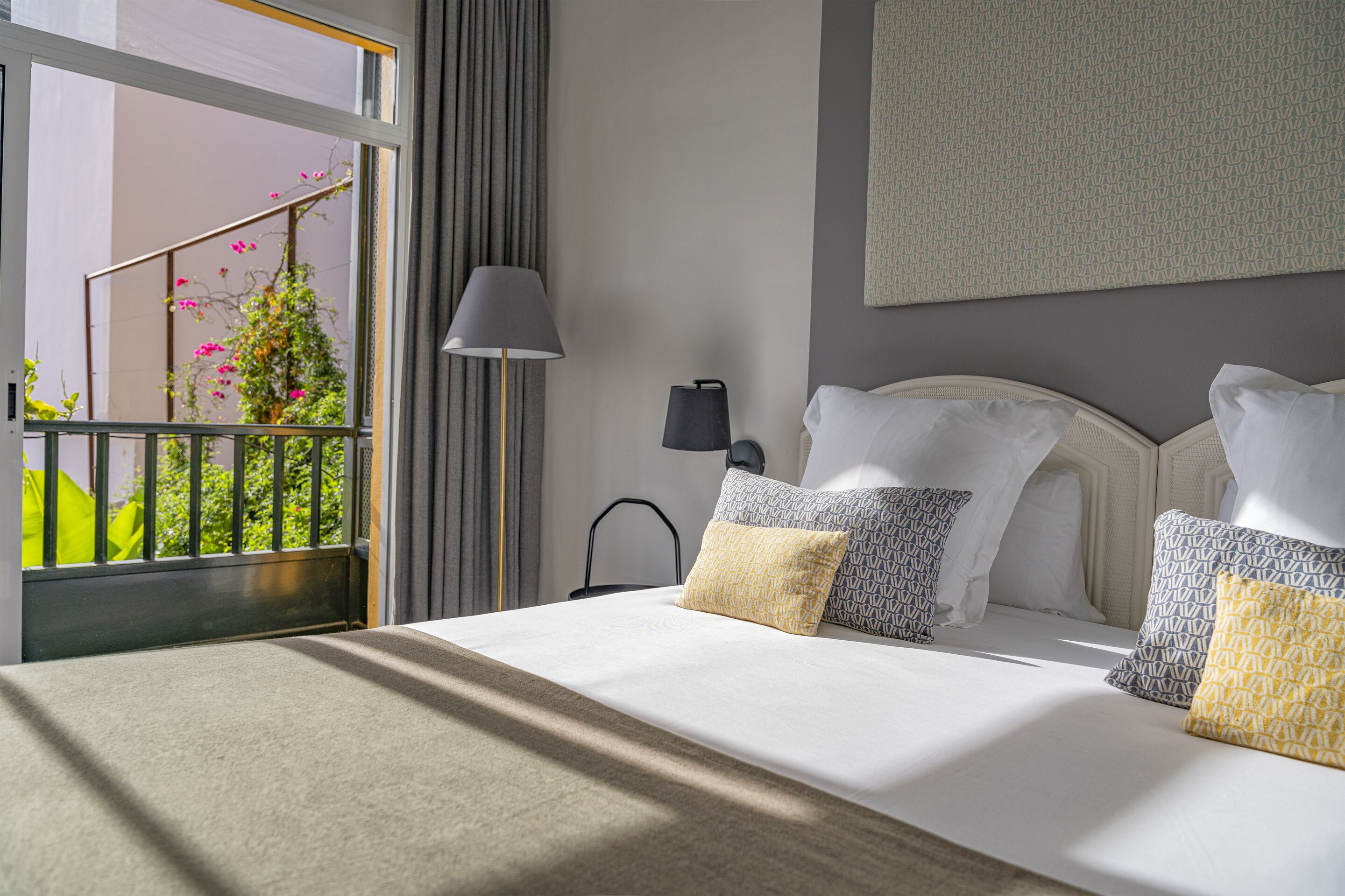 A hotel room with a bed, a nightstand, a lamp, and a view of a balcony with greenery.