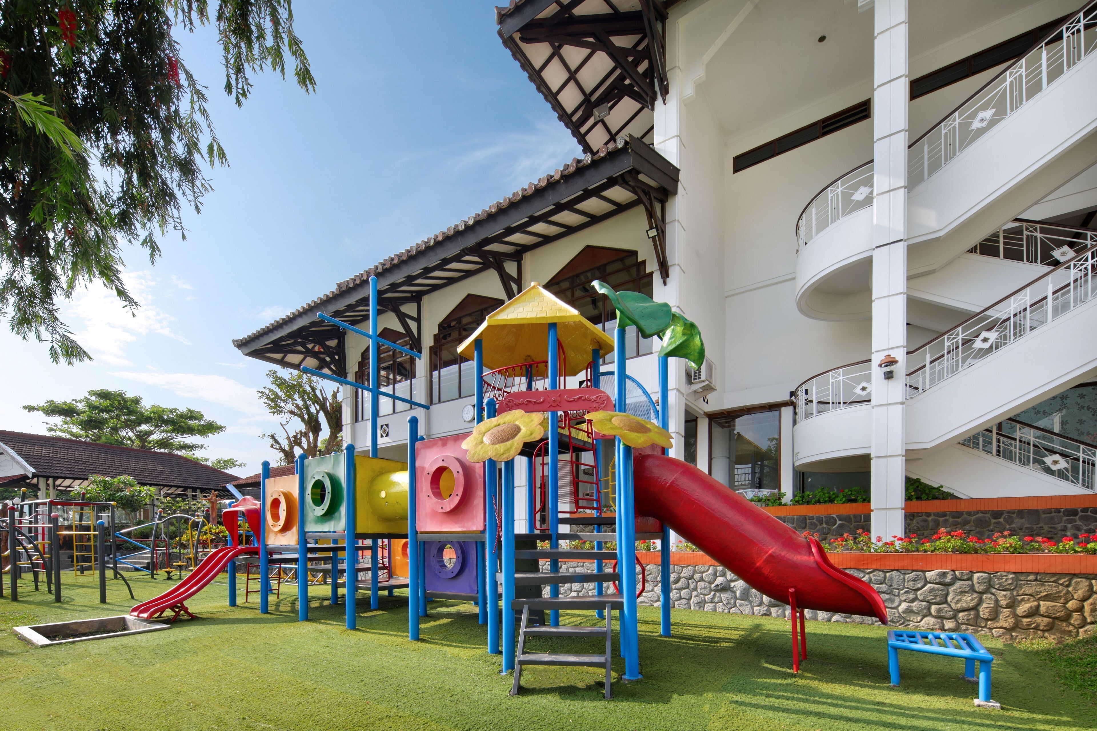 A playground with slides and climbing structures, surrounded by a building with balconies and a stone wall.