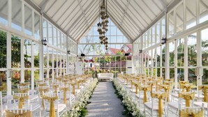 An indoor venue with a glass ceiling, rows of white chairs with gold sashes, and a central aisle leading to a sofa. - Royal Hotel and Villa Batu (Batu)