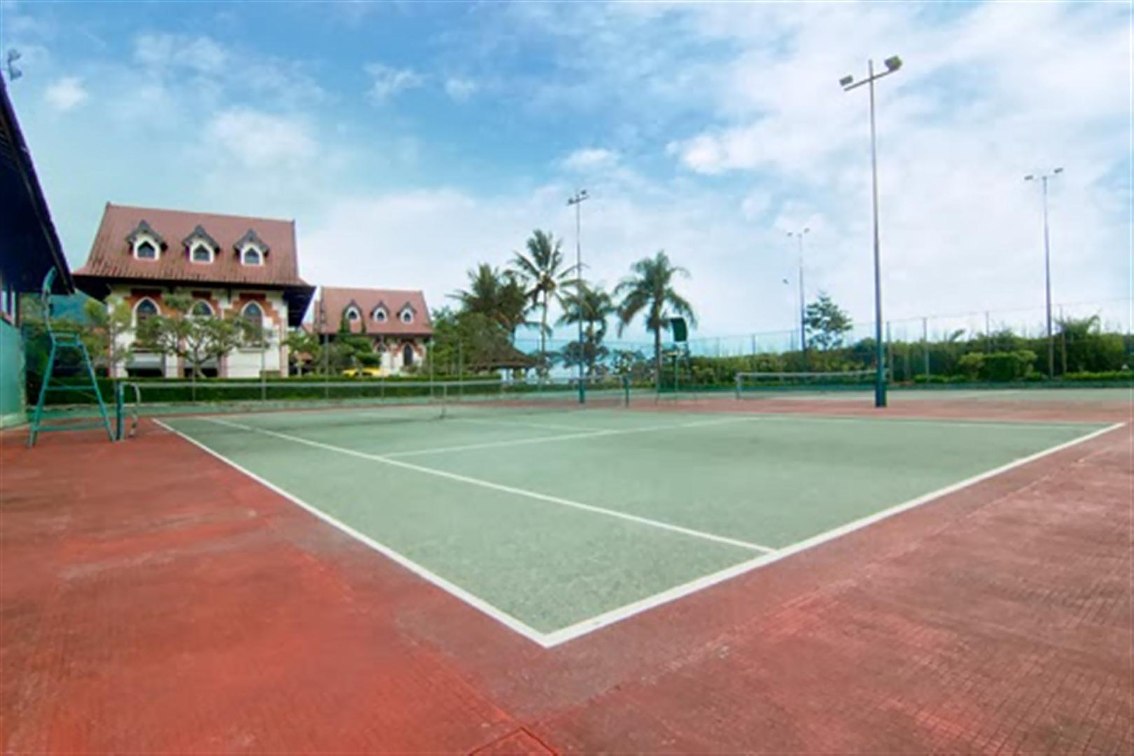An outdoor tennis court with a red surface, surrounded by a fence and palm trees.