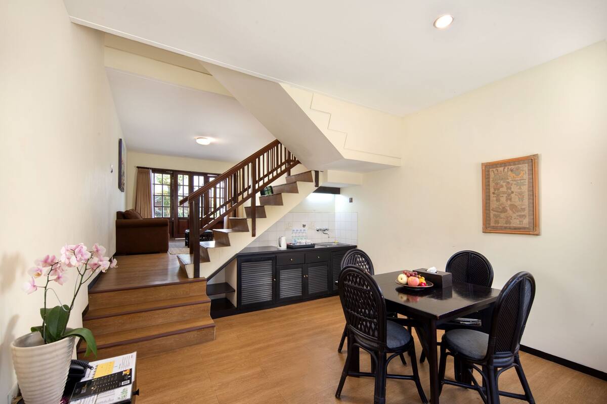 Un vaste espace de vie agrémenté d’un escalier en bois menant à une cuisine, une table à manger avec des chaises et un pot d’orchidée.