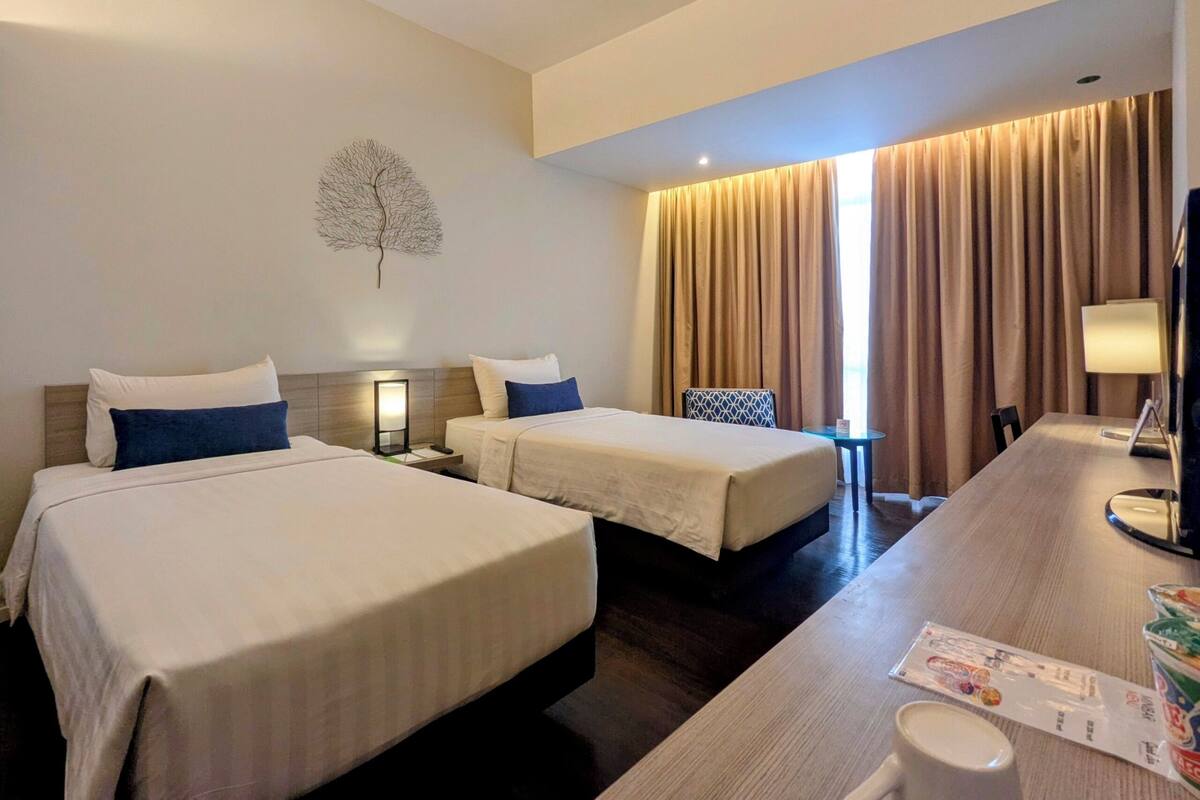 A hotel room with two beds, a desk, a chair, a lamp, and a window with curtains.