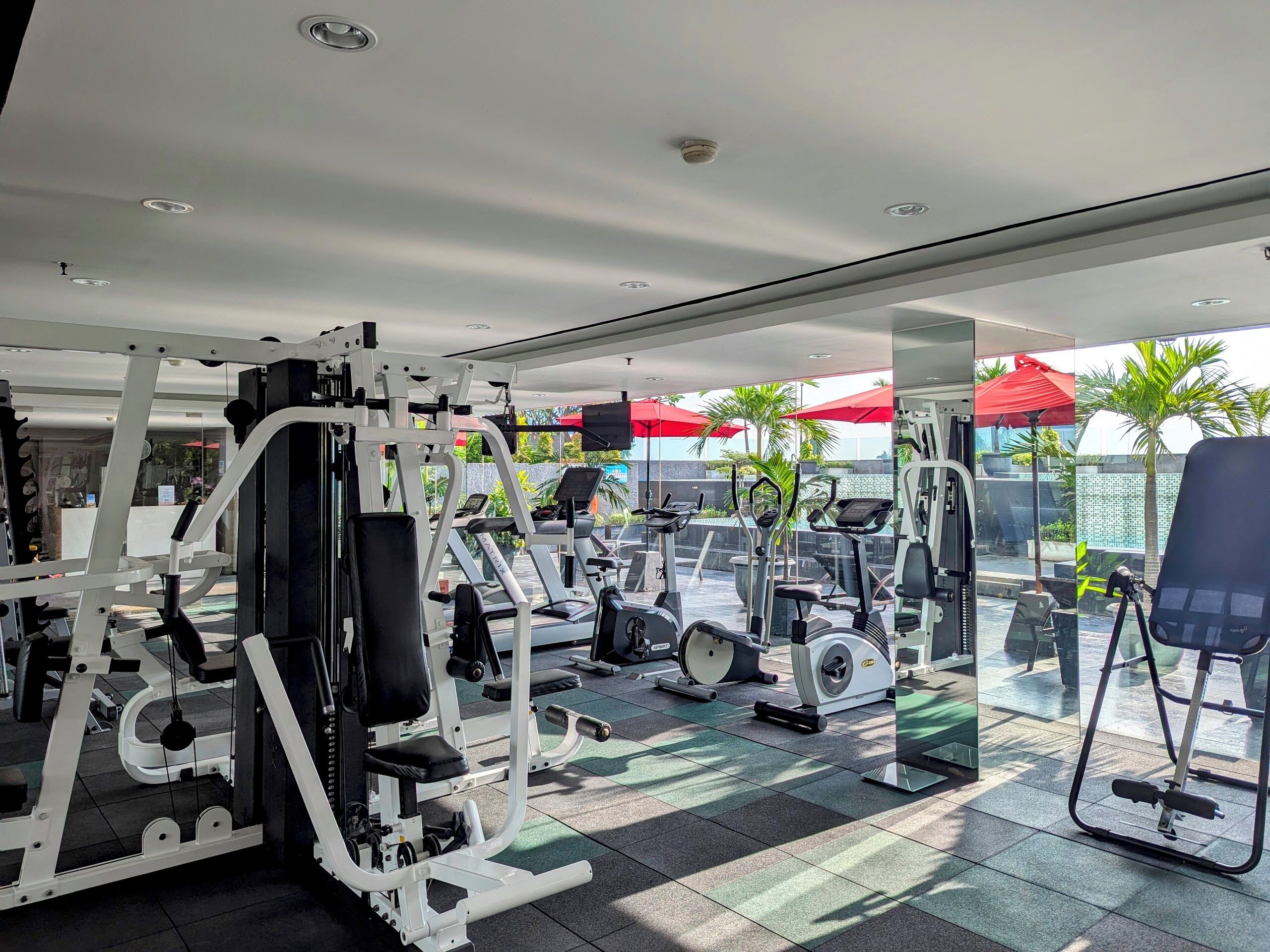 Un gimnasio con máquinas de ejercicio variadas, un espejo grande y zona de estar exterior con sombrillas.