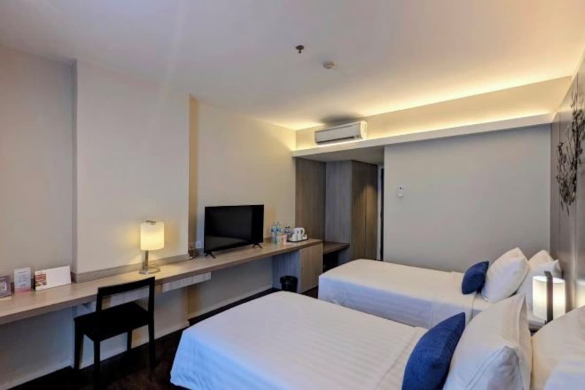 Habitación de hotel con dos camas, un escritorio con televisor, una lámpara y una mesita con tetera.
