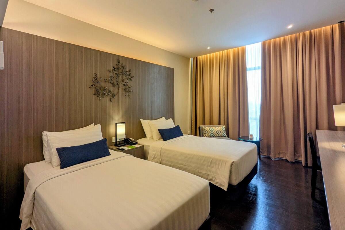A hotel room with two beds, a wooden accent wall, a small table, and a window with curtains.