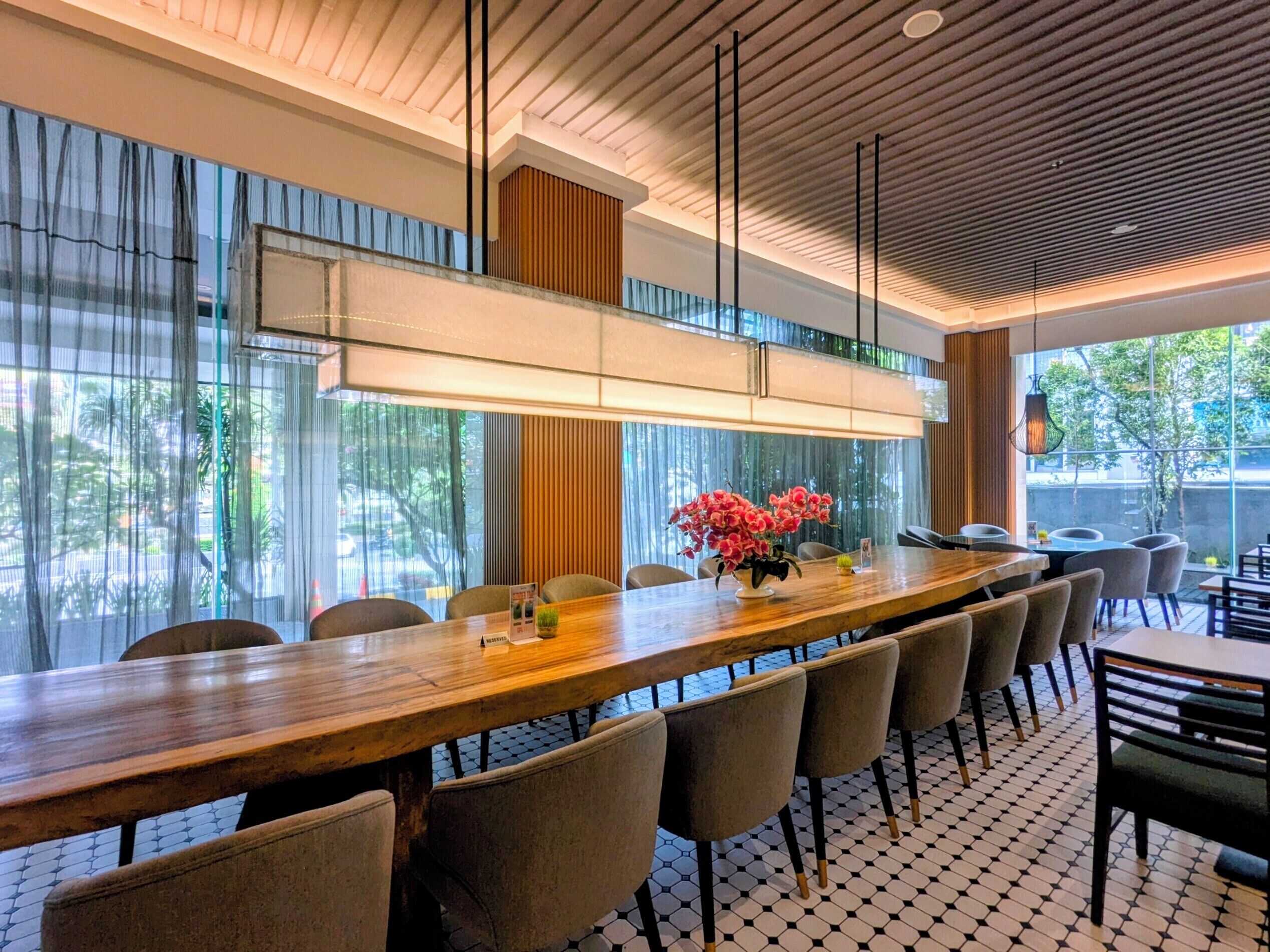A modern dining area with a long wooden table, multiple chairs, and a large window with sheer curtains.
