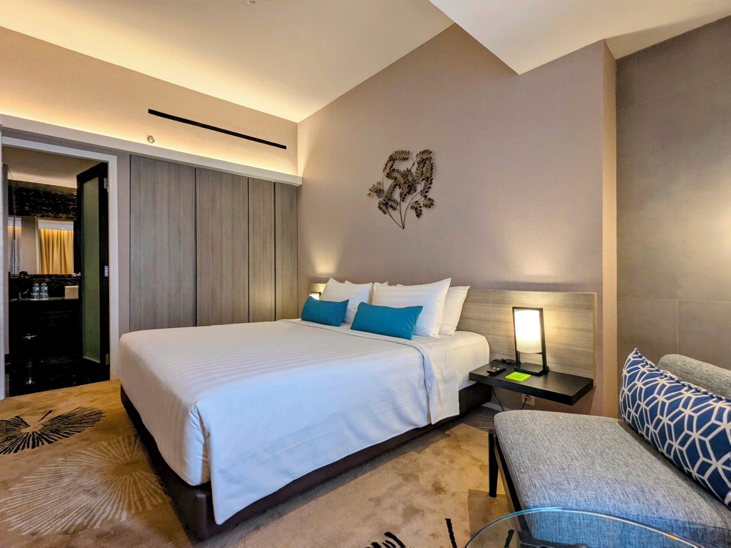 Una habitación de hotel moderna con una cama grande, una mesita de noche con lámpara, una zona de estar con un cojín estampado y un adorno en la pared.