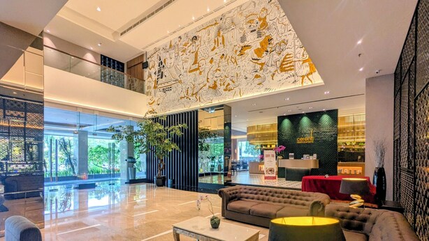 Een moderne hotel lobby met een hoge plafond, een groot muurschildering, een receptiebalie en een zithoek met een bank en salontafel.