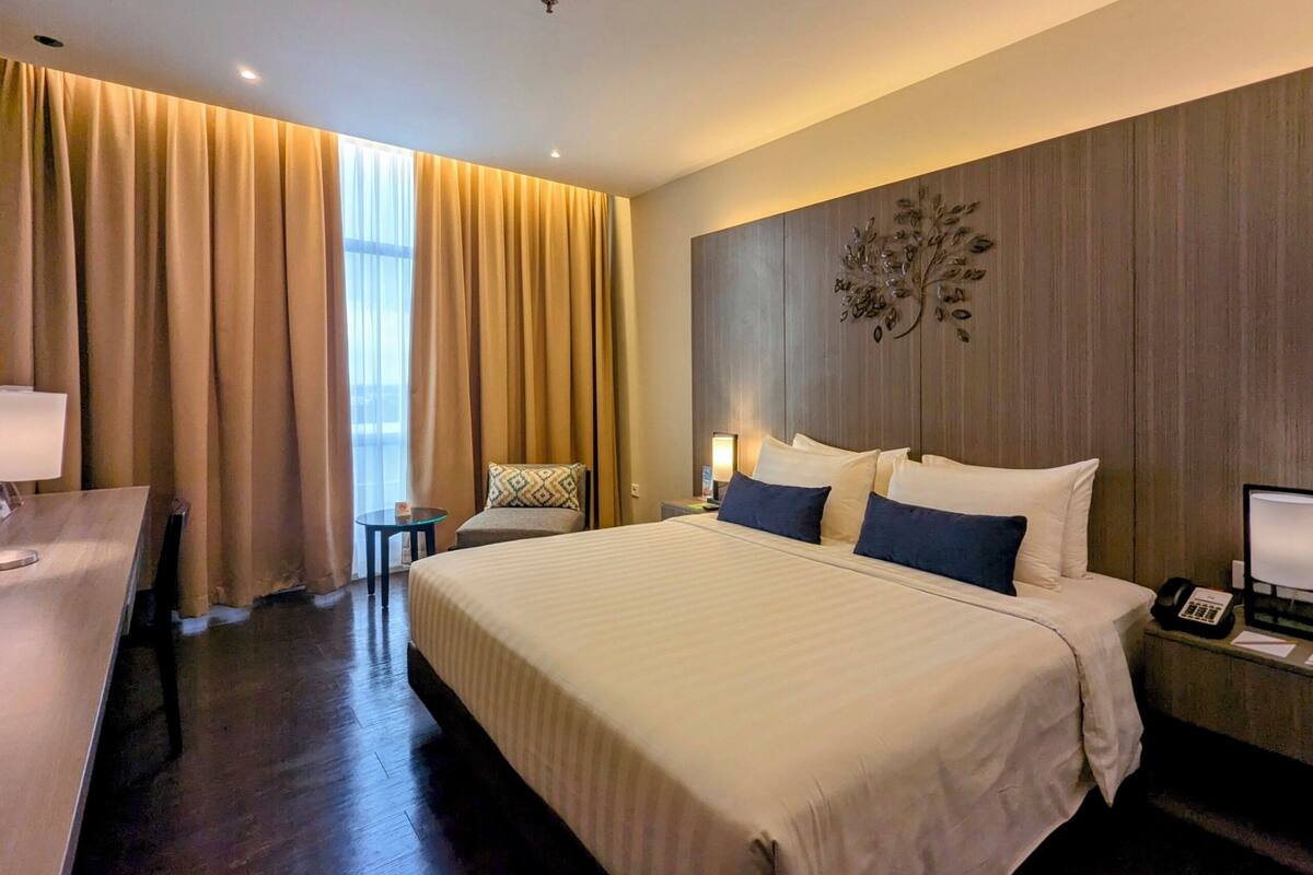A hotel room with a large bed, a desk, a chair, a nightstand with a lamp, and a wall-mounted phone.