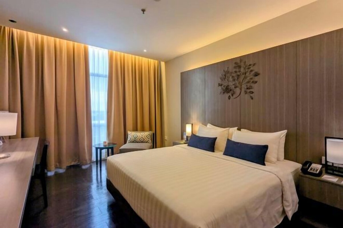 Ein Hotelzimmer mit einem großen Bett, einem Schreibtisch und einem Sessel. Ein Fenster mit Vorhängen, ein Wandbild und ein Nachttisch mit Lampe.