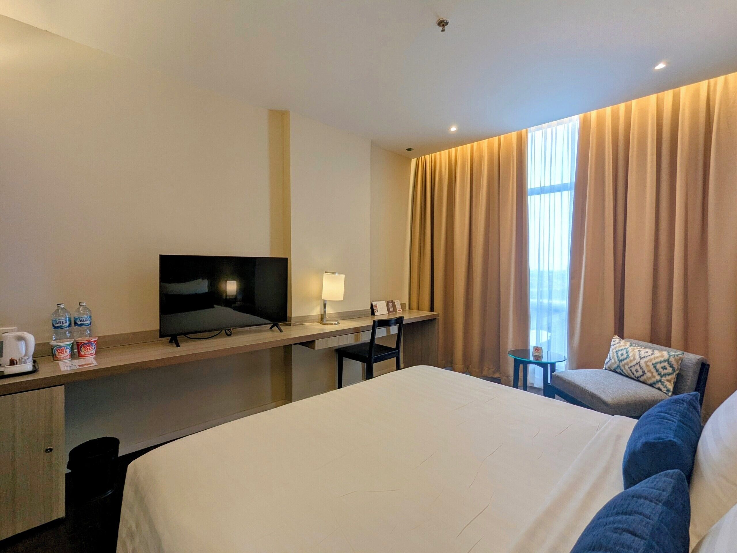 Une chambre d’hôtel comprenant un lit, un bureau avec une télévision, une chaise, une petite table et une fenêtre avec des rideaux.