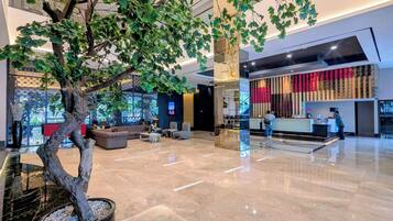 Un lobby moderno con un árbol artificial grande, asientos cómodos, una recepción y una pared decorativa.