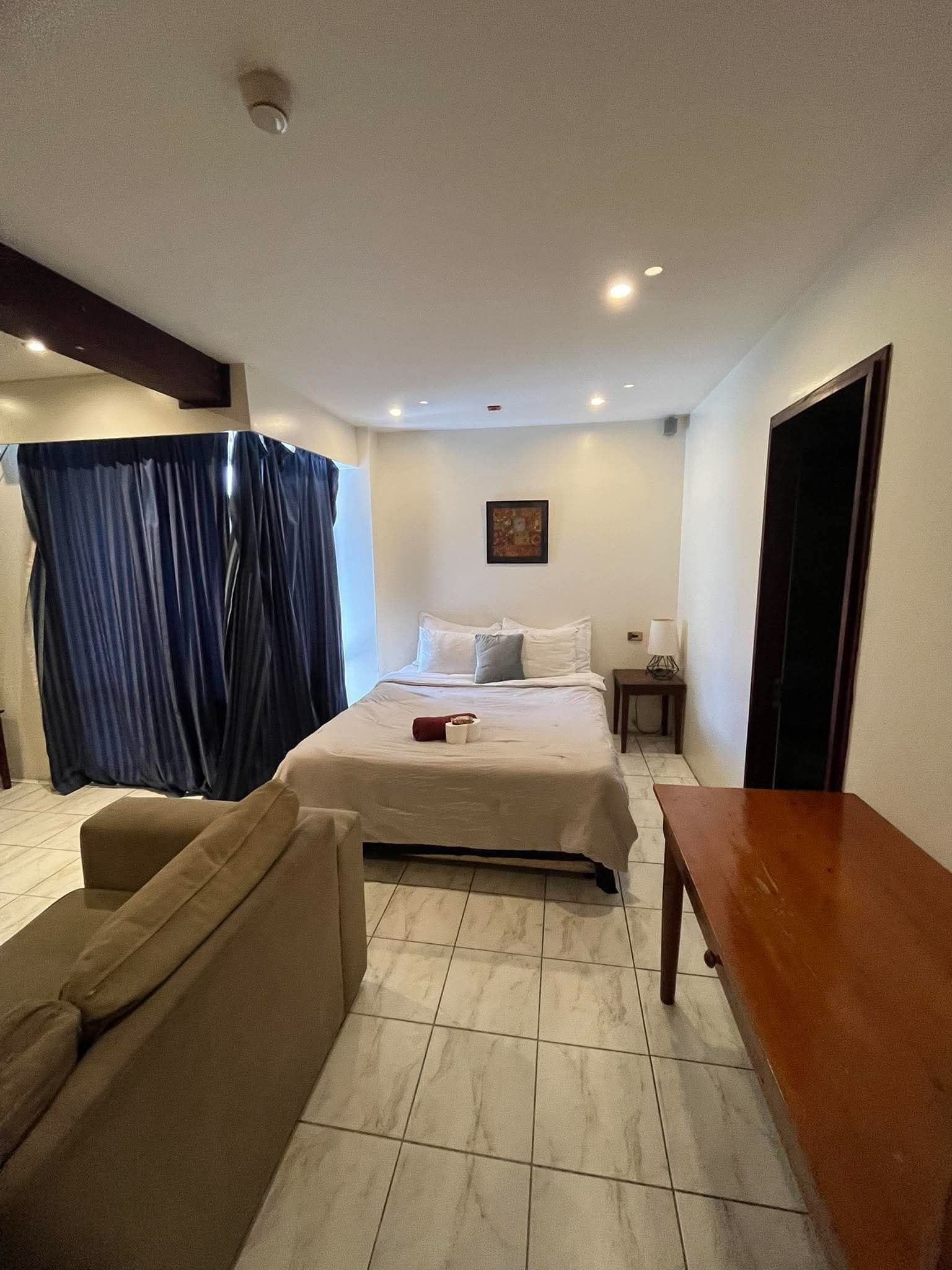 Habitación de hotel con cama, una mesita, una silla, una mesita de noche y una ventana con cortinas.