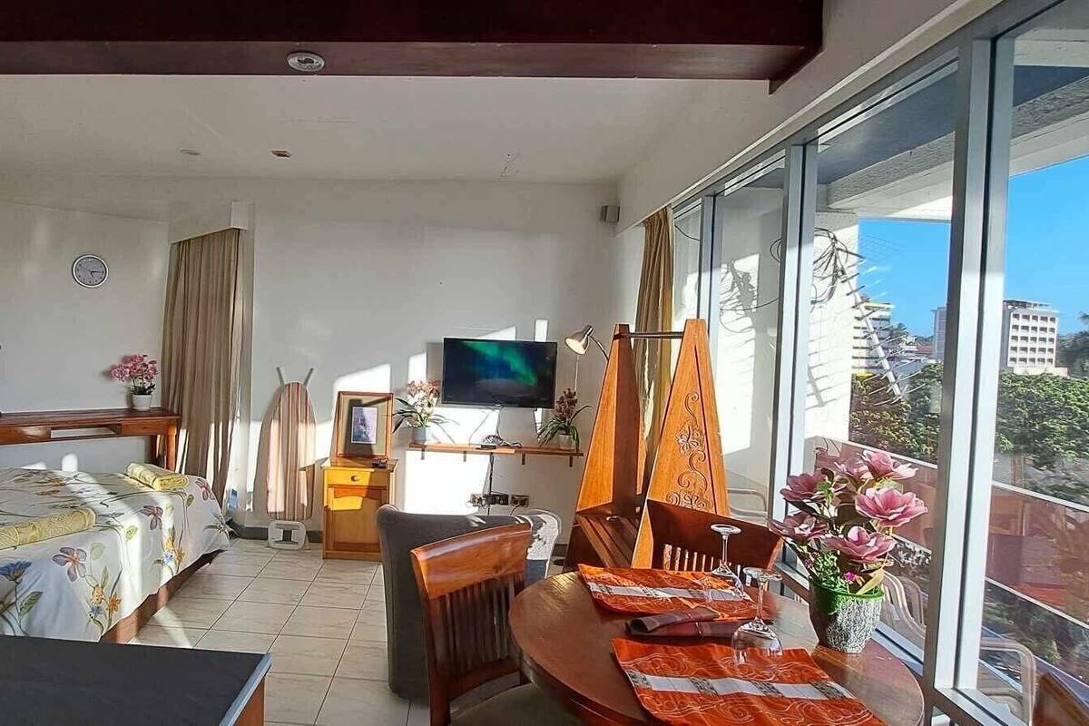 A bedroom with a bed, a desk, a TV, and a balcony with a view of buildings.