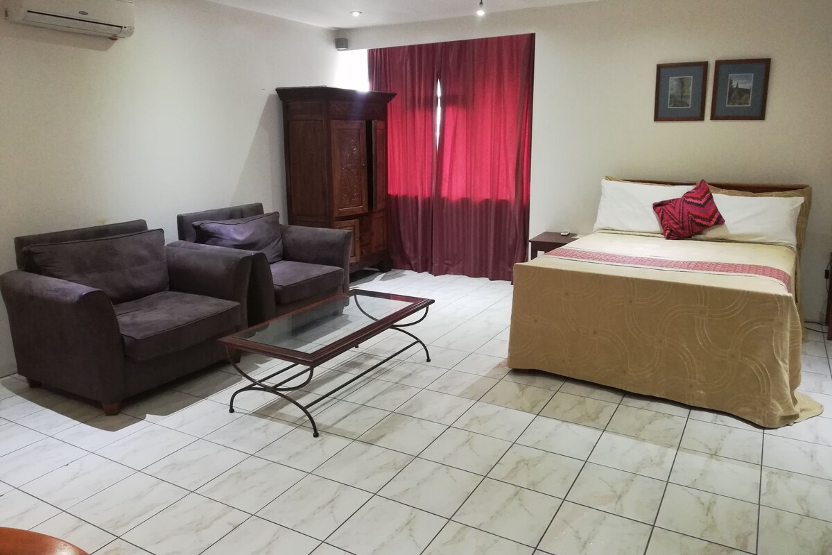 A hotel room with a bed, two armchairs, a small table, a wooden cabinet, and a window with red curtains.