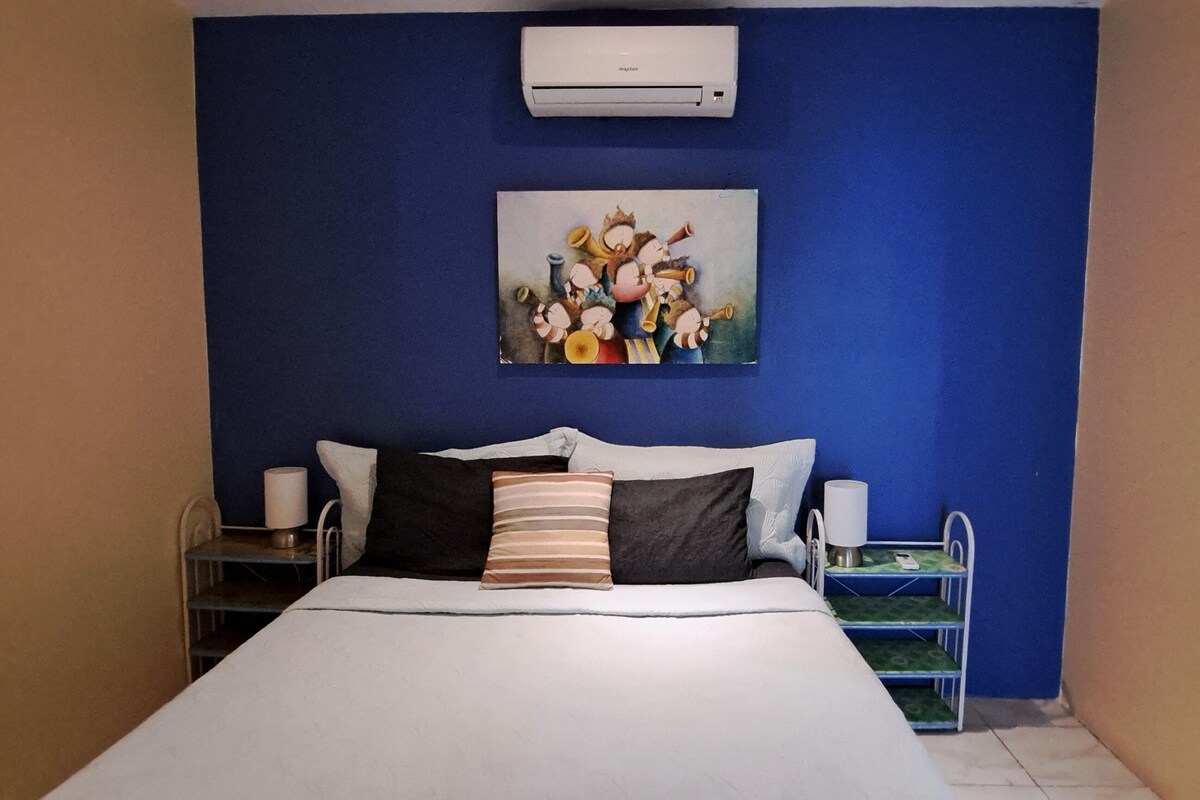 Un dormitorio con cama, almohadas, una mesita de noche, lámpara y aire acondicionado de pared.