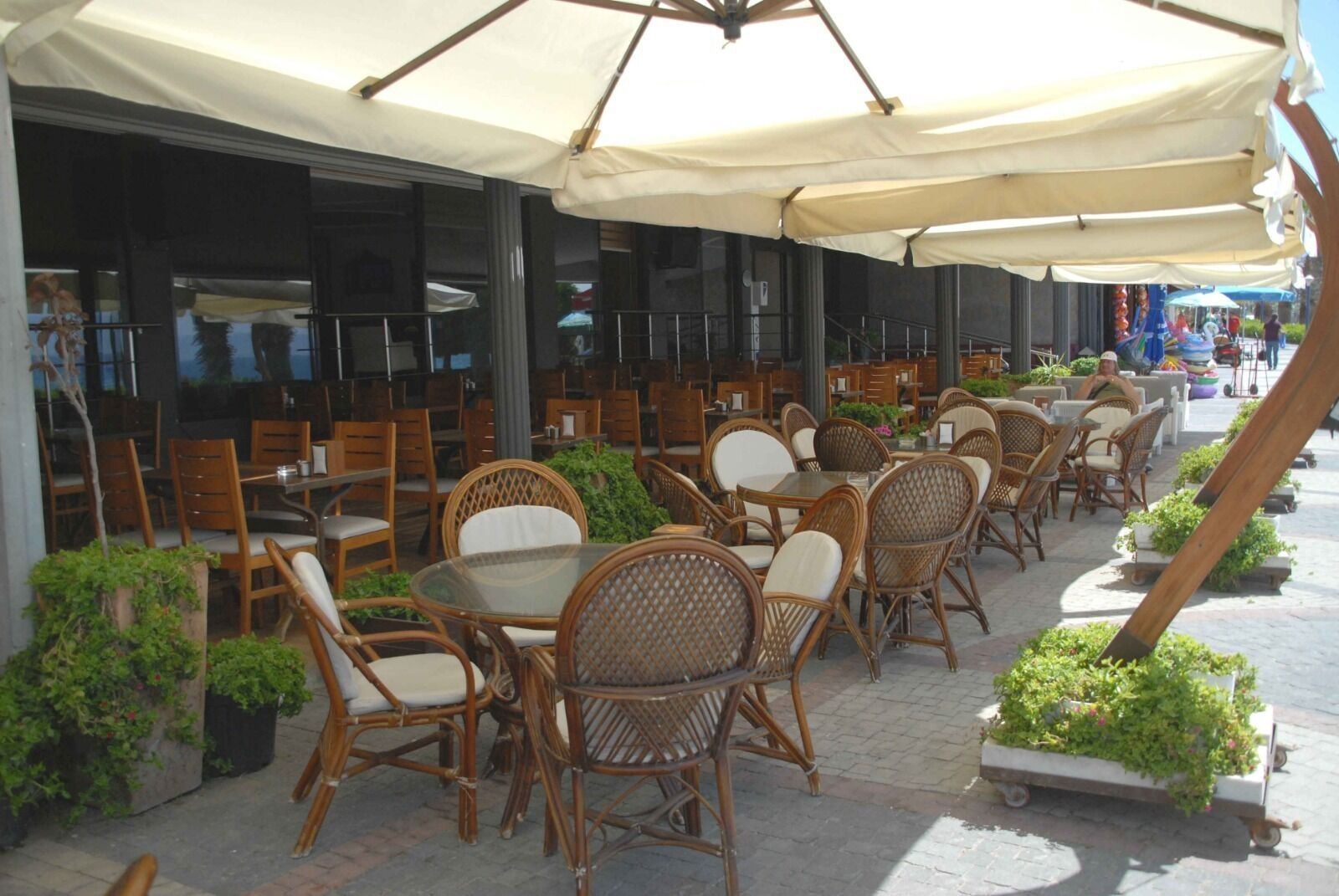 An outdoor cafe with wicker chairs and tables under large umbrellas, surrounded by potted plants.