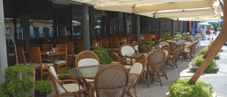 An outdoor cafe with wicker chairs and tables under large umbrellas, surrounded by potted plants.
