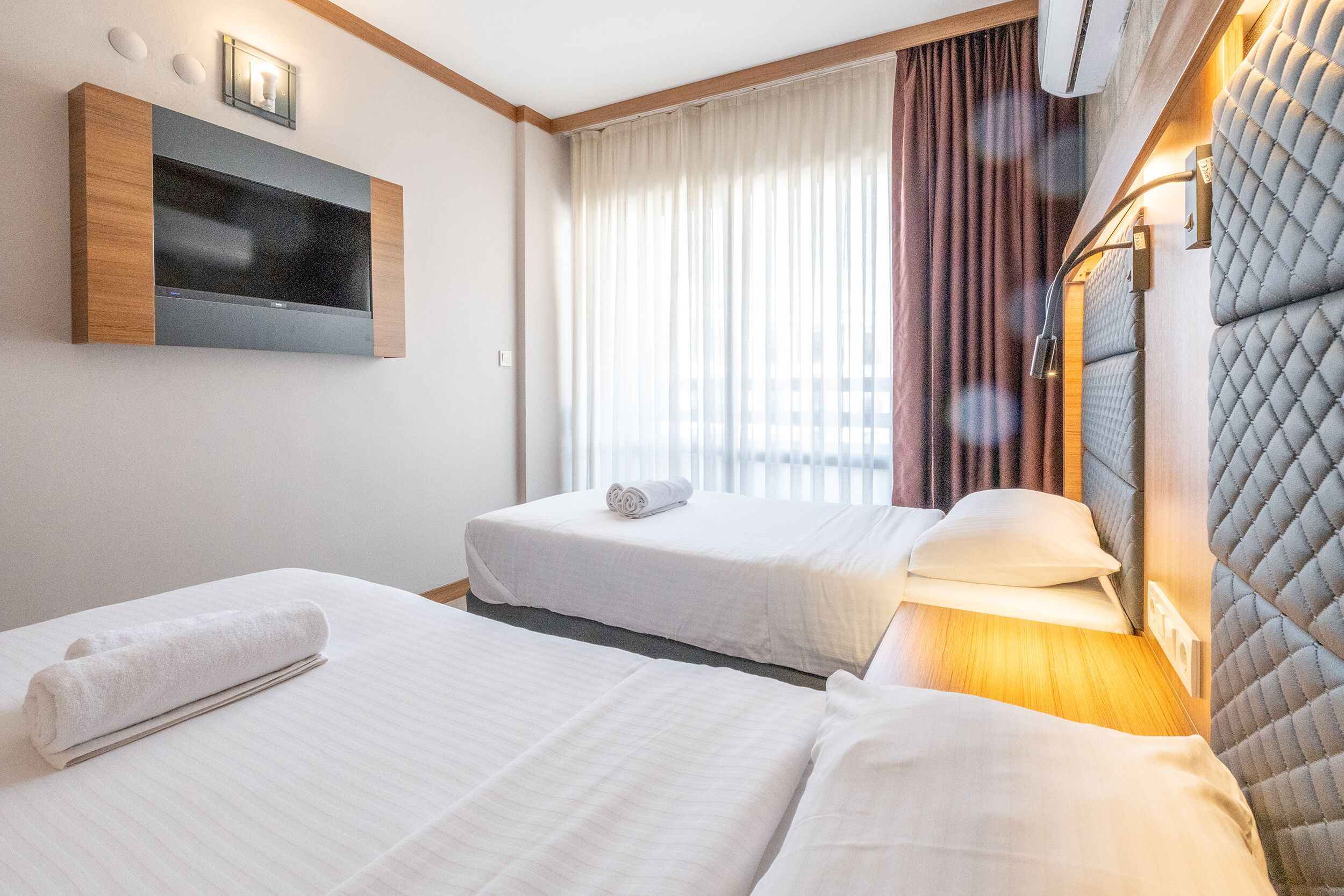 Habitación de hotel con dos camas, un televisor montado en la pared, una ventana con cortinas y un cabecero con iluminación integrada.