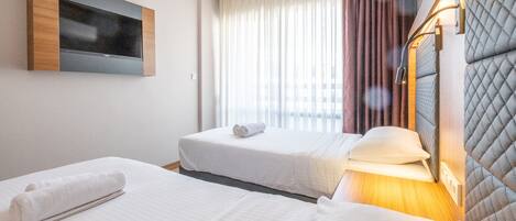 Habitación de hotel con dos camas, un televisor montado en la pared, una ventana con cortinas y un cabecero con iluminación integrada.