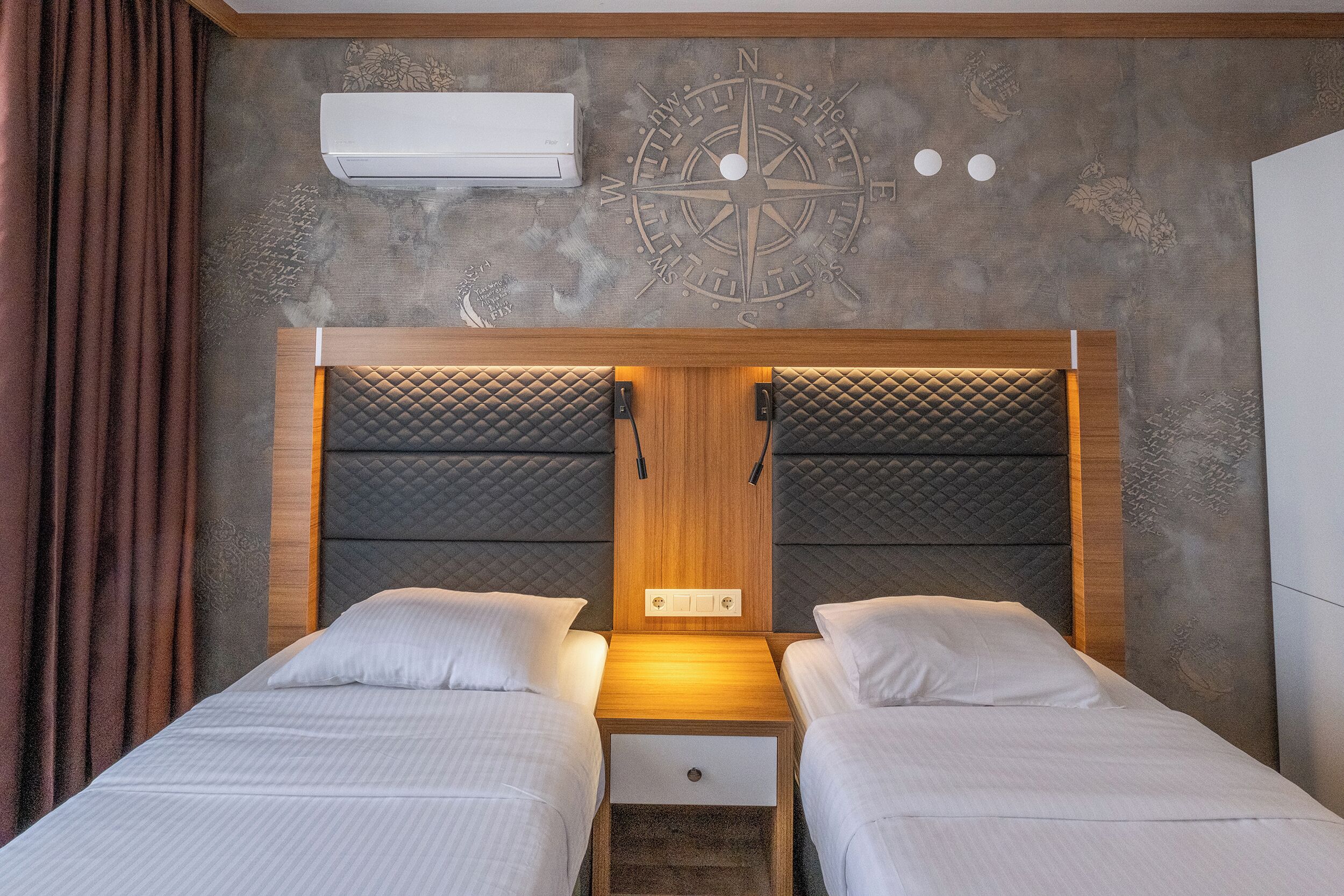 A double bed with a wooden headboard and a small drawer, a compass wall decoration, and an air conditioning unit above the bed.