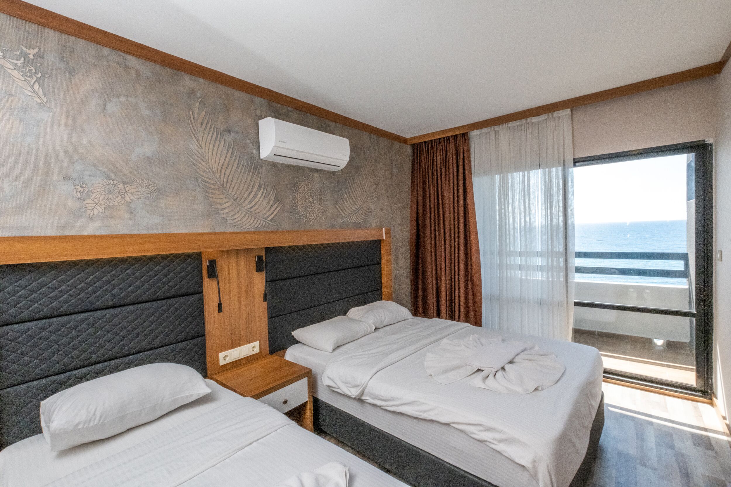 A hotel room with two beds, a wooden headboard, an air conditioning unit, a nightstand, and a view of the sea through a window with curtains.
