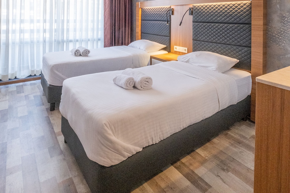 A hotel room with two beds, a large window with sheer curtains, a wall-mounted air conditioner, and a decorative compass design.