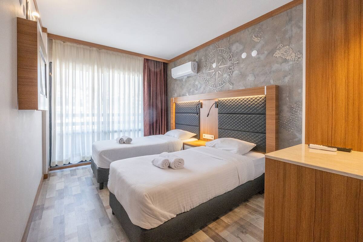 A hotel room with two beds, a wooden headboard, a wall-mounted air conditioner, and a large window with curtains.