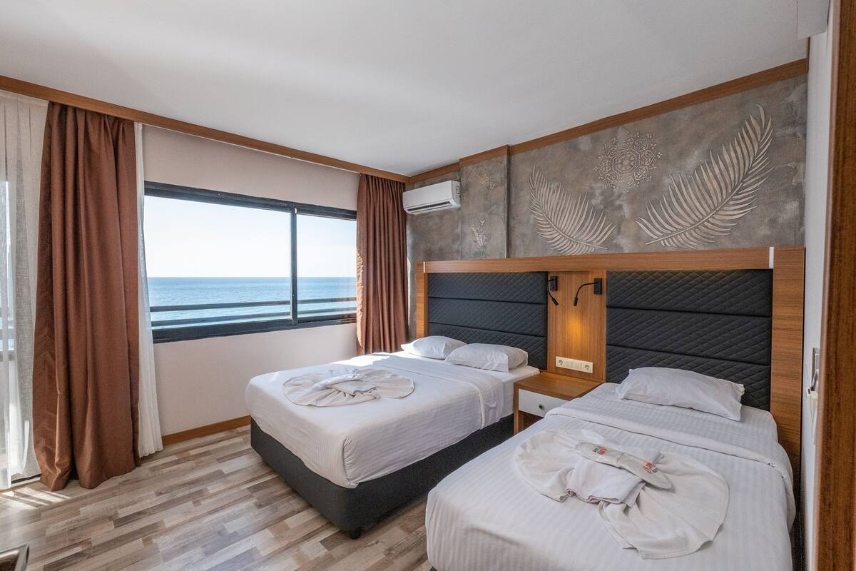 A hotel room with two beds, a large window with curtains, a wall-mounted air conditioner, and a decorative leaf pattern on the wall.