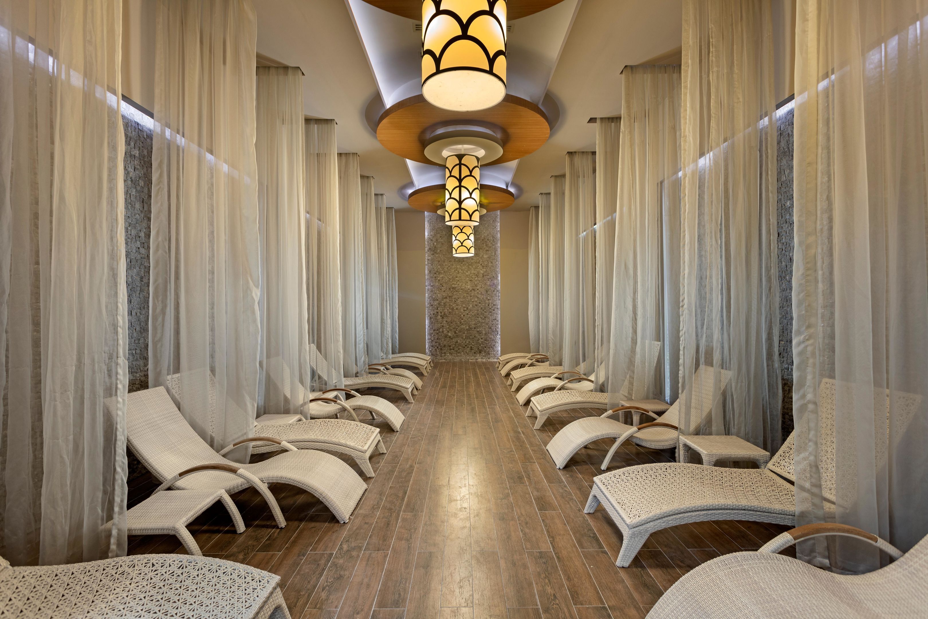 A spa room with rows of lounge chairs, a central light fixture, and a stone wall in the background.
