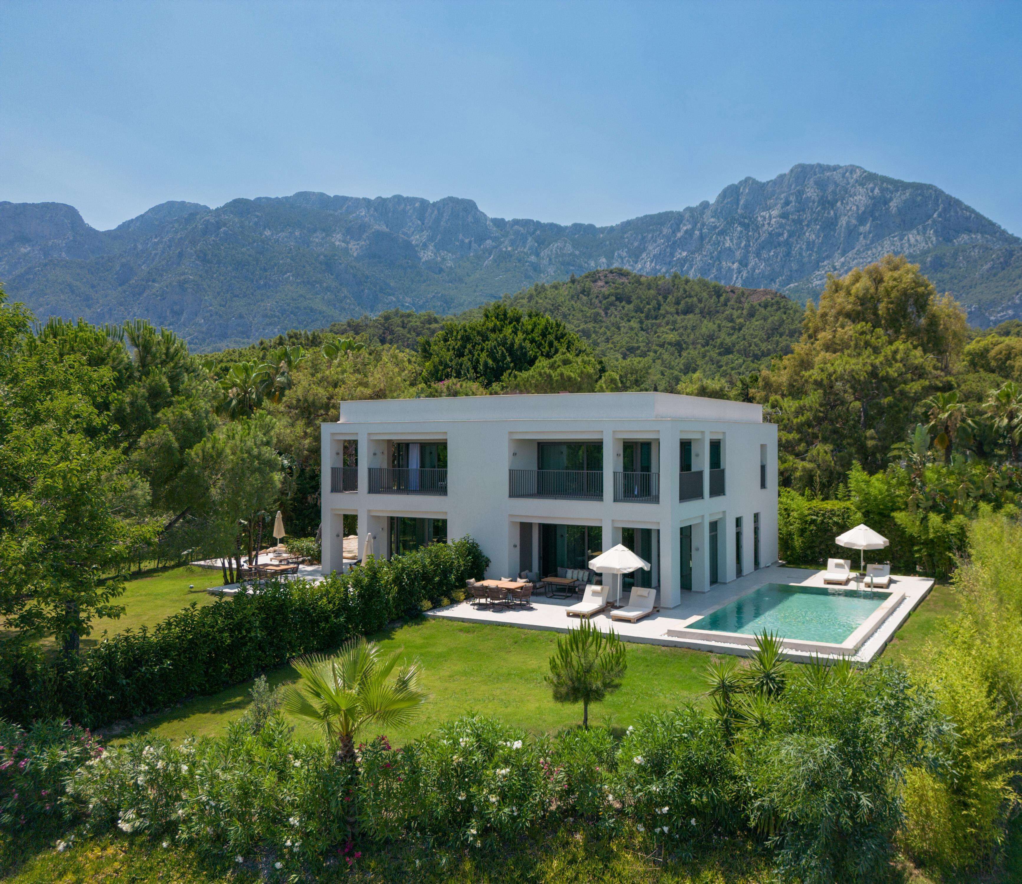 A modern two-story house with a swimming pool, surrounded by lush greenery and mountains.